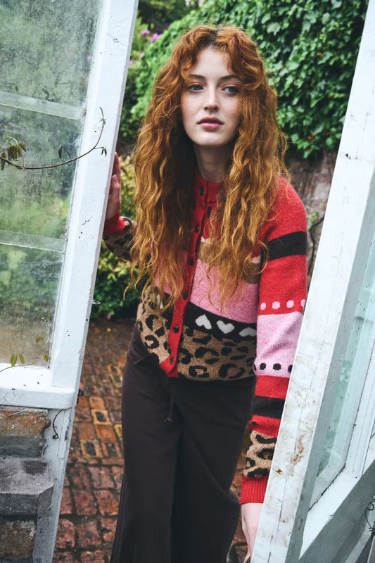 A woman with long, wavy red hair stands in an open white greenhouse doorway, wearing a color-blocked knit cardigan with leopard print, pink, red, and black panels, paired with dark brown pants.