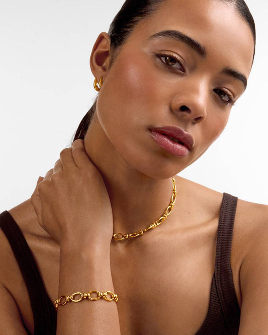A close-up of a model wearing a gold chain necklace, matching gold hoop earring, and gold link bracelet against a light gray background.