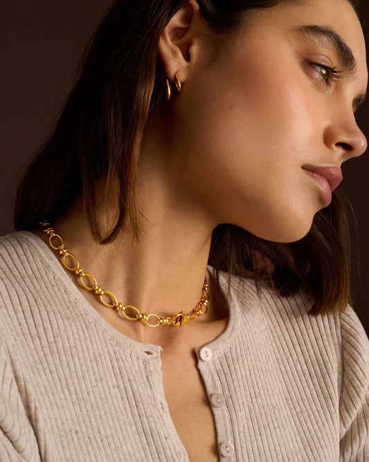 A close-up side profile of a woman wearing a chunky gold chain necklace with oval links and a small gold hoop earring, paired with a ribbed beige top against a dark brown background.