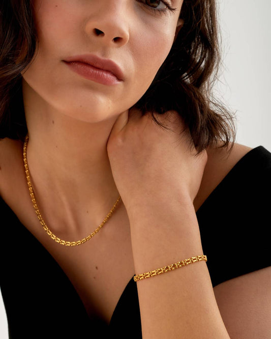 A close-up lifestyle shot of a woman wearing a delicate gold chain necklace and a matching gold link bracelet, styled with a black top against a light gray background.