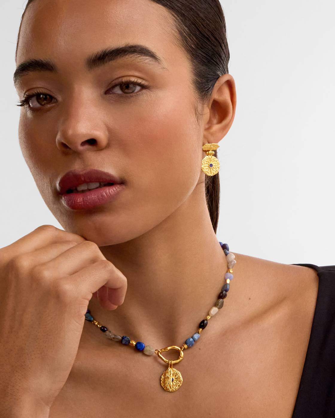 A close-up of a model wearing a gold pendant necklace with blue and beige beads, and a matching gold drop earring, against a light gray background.