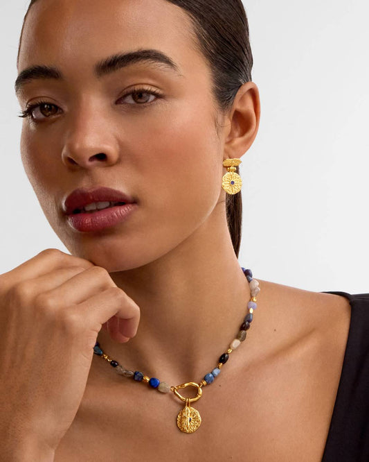 A close-up of a model wearing a gold pendant necklace with blue and beige beads, and a matching gold drop earring, against a light gray background.