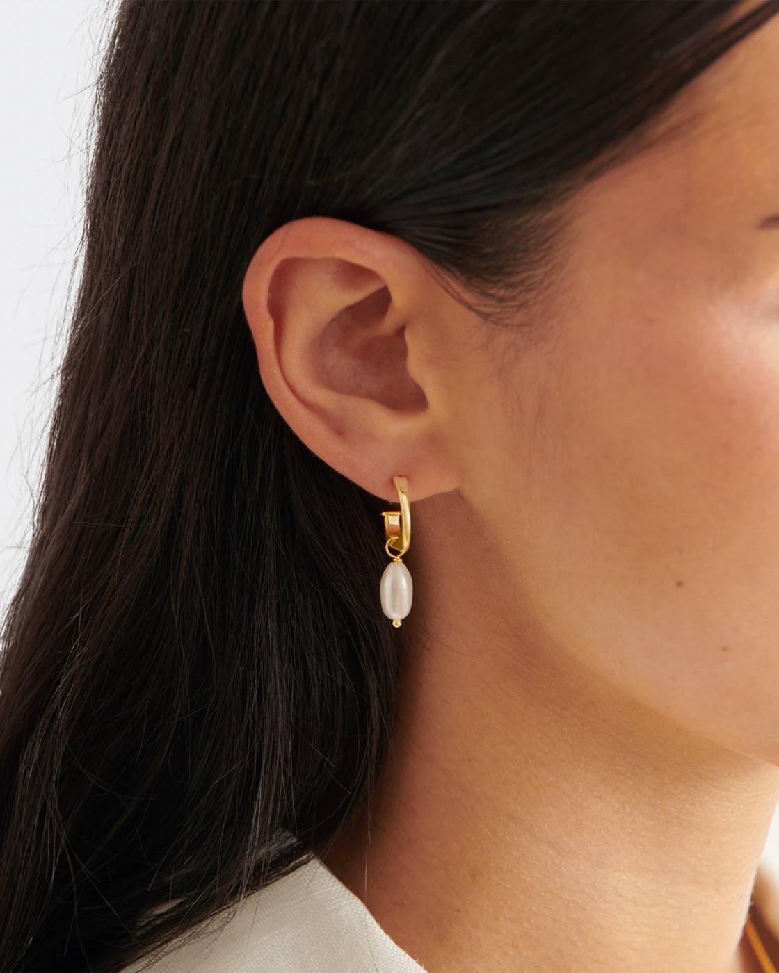 A close-up of a model's ear wearing a delicate gold huggie hoop earring with a small white pearl drop.