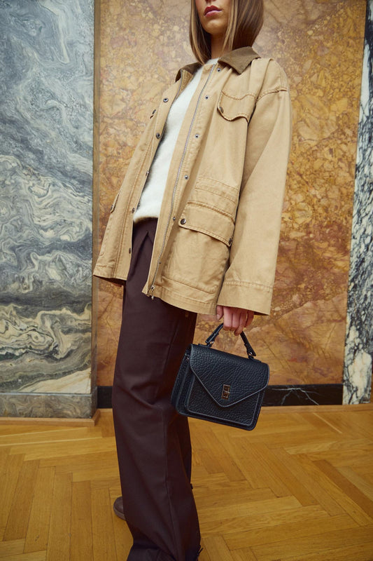 Person wearing a beige jacket and holding a black handbag against a marble wall.