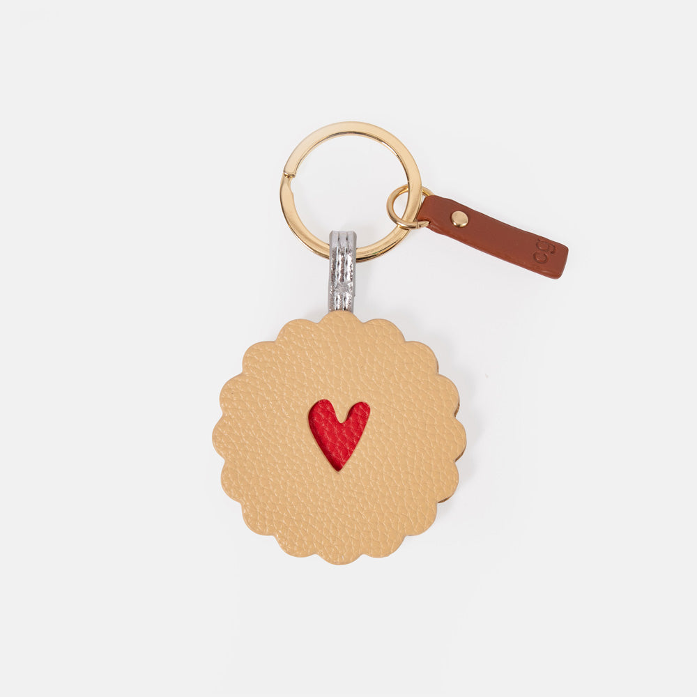 A round, scalloped-edge leather keychain charm designed like a sandwich cookie, featuring a textured tan body with a small red heart in the center. It is attached to a gold-tone split ring and a brown leather tag with a gold rivet, set against a plain light gray background.