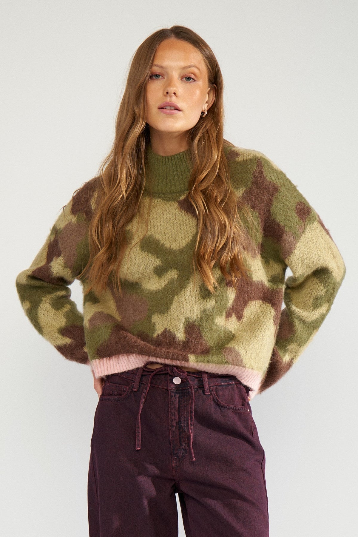 Numph Camu pullover in Calla Green = A woman modeling a fuzzy camouflage knit sweater with olive green, brown, and beige tones, paired with dark purple pants, against a plain light grey background.