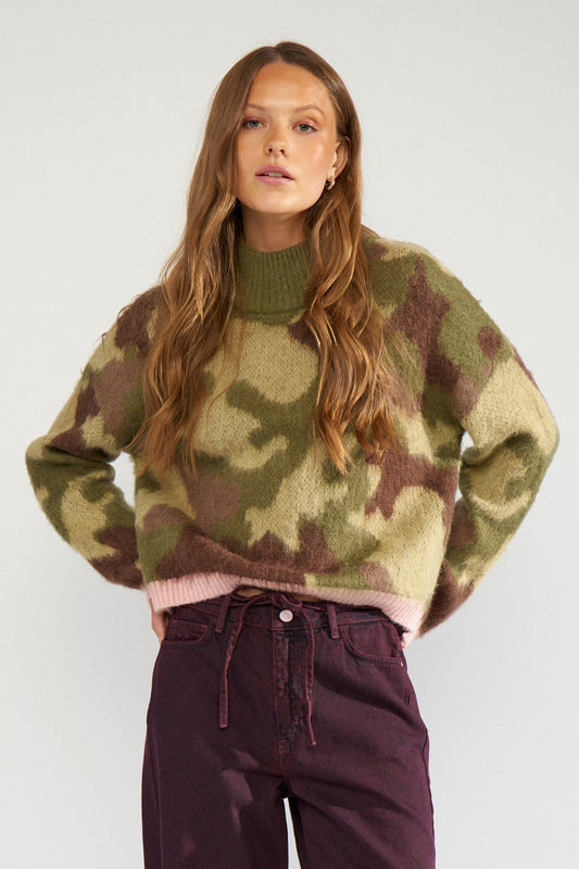 Numph Camu pullover in Calla Green = A woman modeling a fuzzy camouflage knit sweater with olive green, brown, and beige tones, paired with dark purple pants, against a plain light grey background.