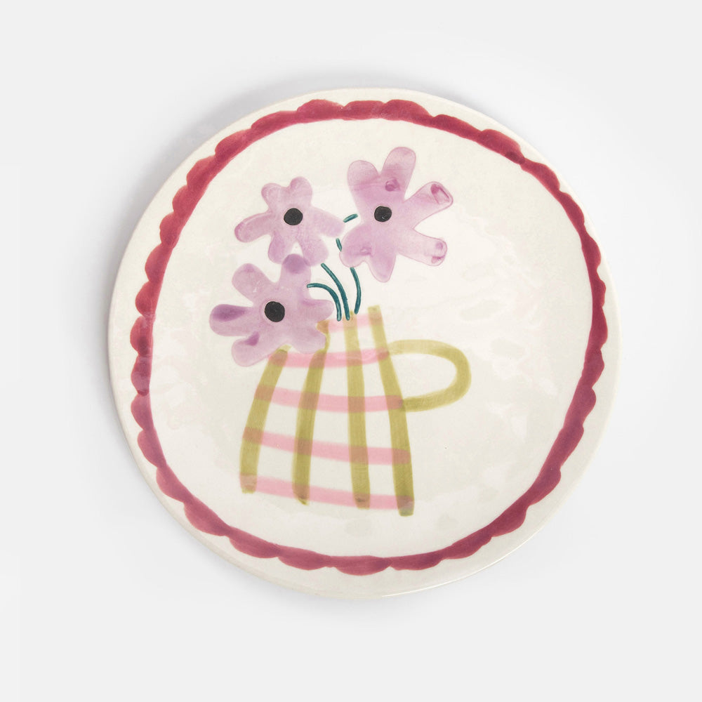 A round ceramic plate with a scalloped dark red rim, featuring a hand-painted illustration of pink flowers in a striped yellow-green vase on a cream background.