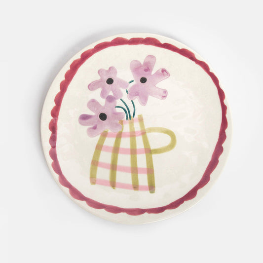 A round ceramic plate with a scalloped dark red rim, featuring a hand-painted illustration of pink flowers in a striped yellow-green vase on a cream background.