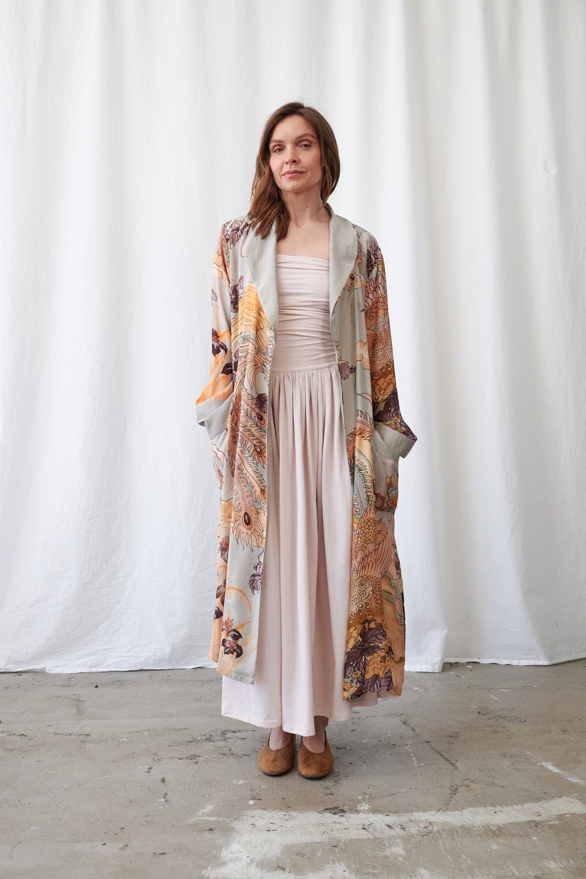 A woman stands in front of a white curtain backdrop wearing a long, light blush pink pleated dress with a matching sheer, lightweight open-front cardigan featuring a warm-toned abstract print in orange, beige, and brown.