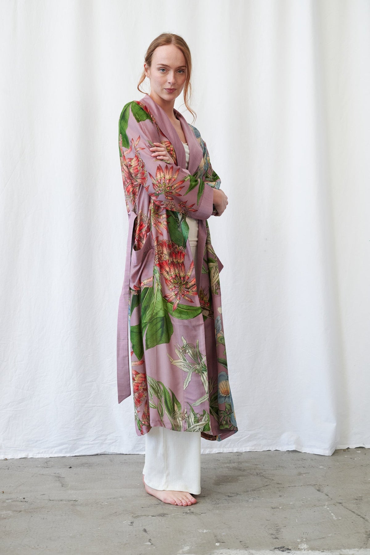 One Hundred Stars Rhodoleia Pink Gown - A woman stands barefoot in front of a white curtain backdrop, wearing a long, loose floral robe or kimono-style wrap in muted mauve with green leaves and red-orange flowers, paired with white wide-leg pants.