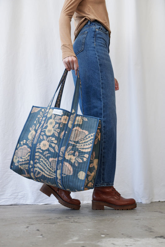 A person wearing blue jeans and brown boots carries a large blue tote bag with a beige floral and paisley print against a white curtain backdrop.