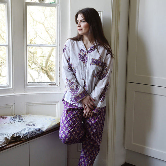 A woman wearing a white pajama set with purple and gold floral patterns leans against a white paneled wall next to a window.