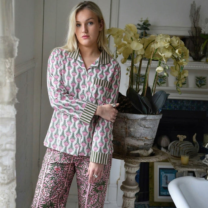 A woman modeling a two-piece pajama set featuring a pink and green floral pattern on a white background, with striped collar and cuffs, standing in a cozy room next to a potted orchid.