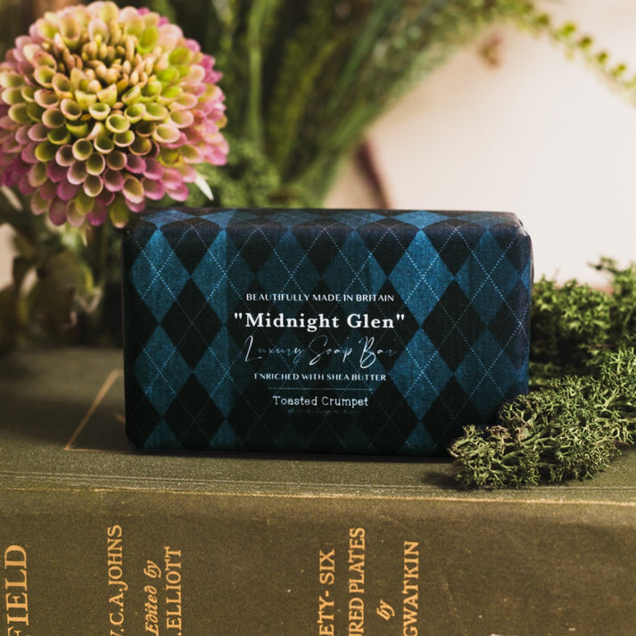 Toasted Crumpet Midnight Glen Soap - A rectangular bar of soap wrapped in a dark blue and black argyle patterned paper sleeve, resting on a vintage green book surrounded by flowers and greenery.