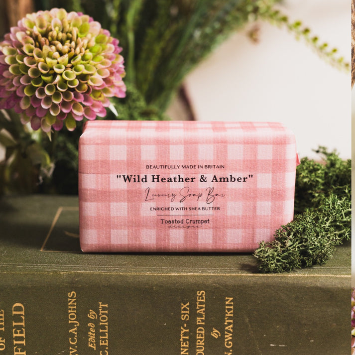 Toasted Crumpet Wild Heather & - Amber Soap =A pink and white gingham wrapped soap bar labeled 'Wild Heather & Amber Luxury Soap Bar' resting on a dark green book, surrounded by fresh greenery and flowers.