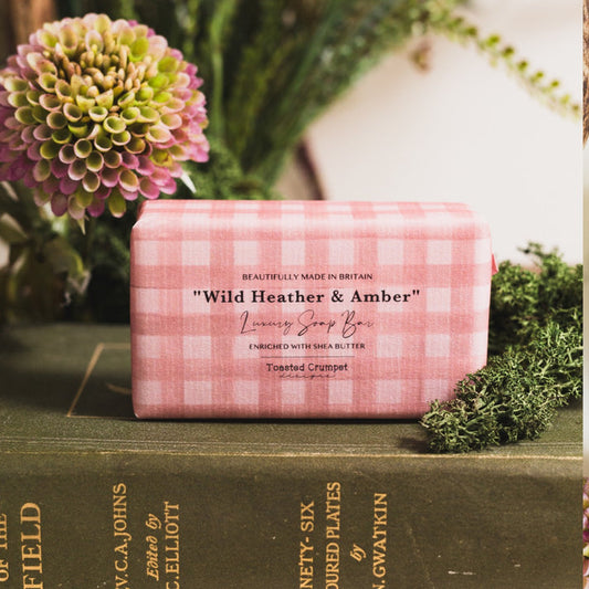 Toasted Crumpet Wild Heather & - Amber Soap =A pink and white gingham wrapped soap bar labeled 'Wild Heather & Amber Luxury Soap Bar' resting on a dark green book, surrounded by fresh greenery and flowers.