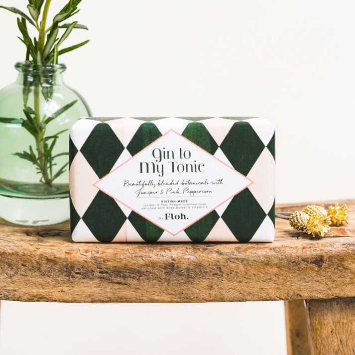 Toasted Crumpet Gin to my Tonic Soap - A rectangular bar of soap wrapped in a dark green and white argyle patterned paper sleeve, labeled 'Gin to My Tonic' by the brand Floh. The soap bar sits on a rustic wooden table next to a glass vase with rosemary and small dried flowers.