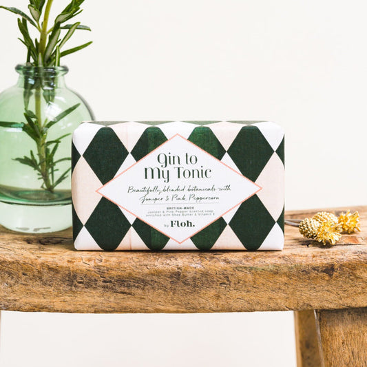 Toasted Crumpet Gin to my Tonic Soap - A rectangular bar of soap wrapped in a dark green and white argyle patterned paper sleeve, labeled 'Gin to My Tonic' by the brand Floh. The soap bar sits on a rustic wooden table next to a glass vase with rosemary and small dried flowers.