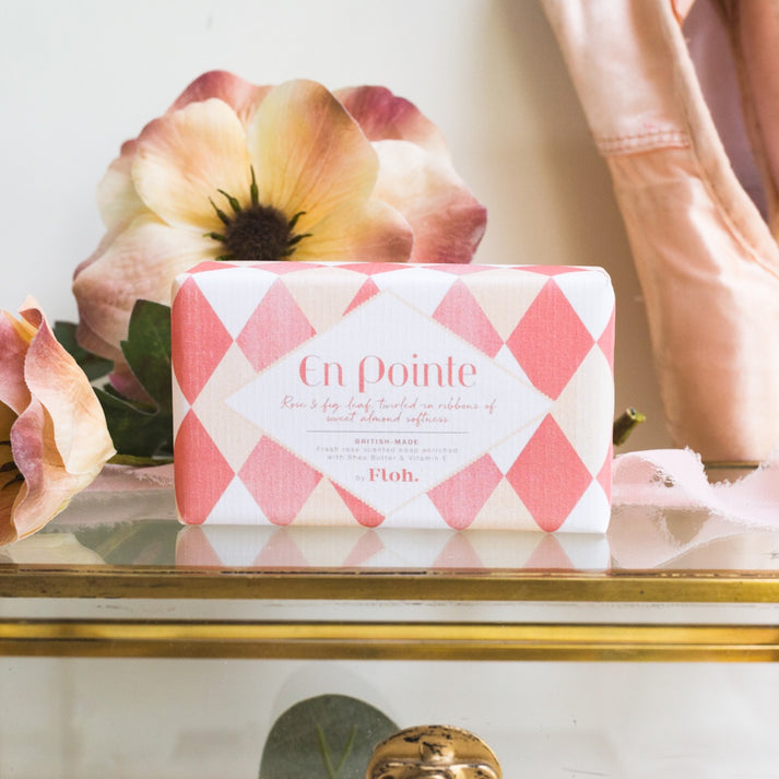 Toasted Crumpet En Pointe Soap - A rectangular bar of En Pointe French Shell soap by Floh, wrapped in a pink and white argyle patterned paper, displayed on a gold-trimmed shelf with soft-focus flowers and a pink garment in the background. 