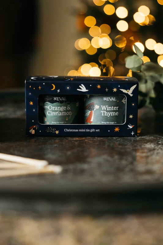 SEVAL Christmas mini tin gift set containing Orange & Cinnamon and Winter Thyme mugs, presented in a dark blue starry gift box on a dark tabletop with warm bokeh lights in the background.