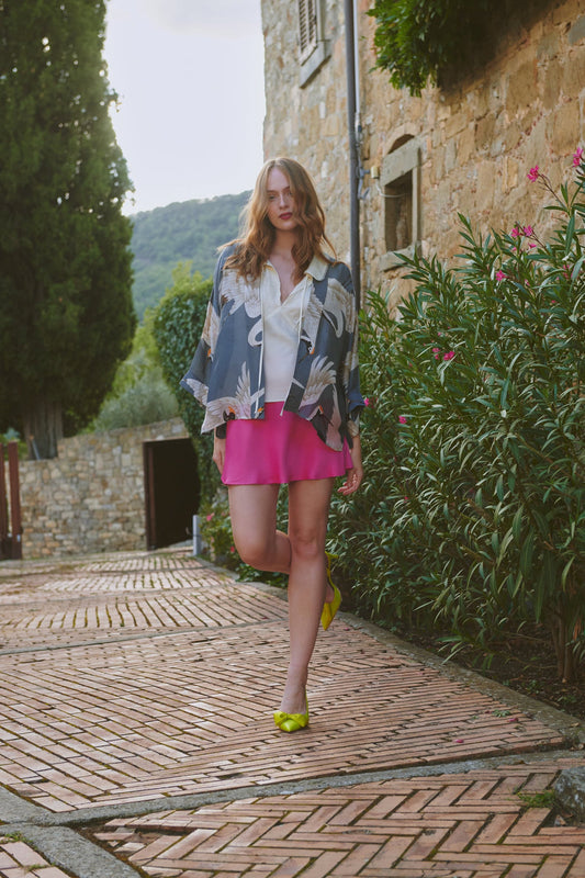 Woman in a grey stork patterned kimono and pink skirt walking on a stone path with greenery and a building in the background.