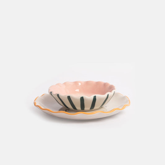A small scalloped ceramic bowl with a pink interior and dark green vertical stripes, placed on a matching shallow plate with a yellow-orange scalloped rim, photographed on a clean white background.