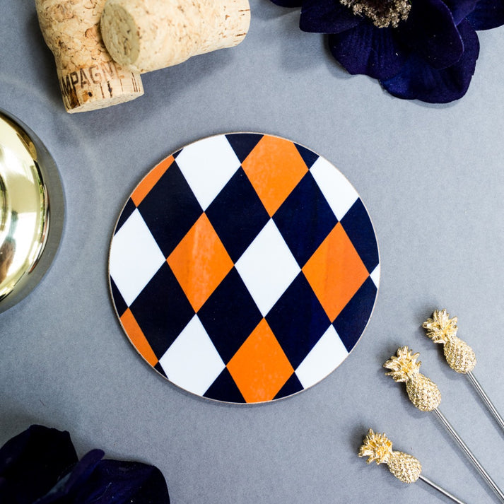 Toasted Crumpet Cobalt & Orche (Harlequin) Round Coaster - A round ceramic coaster with a bold orange, white, and navy blue harlequin diamond pattern, styled on a gray tabletop with decorative props including a cork, a gold bowl, and pineapple-shaped picks.