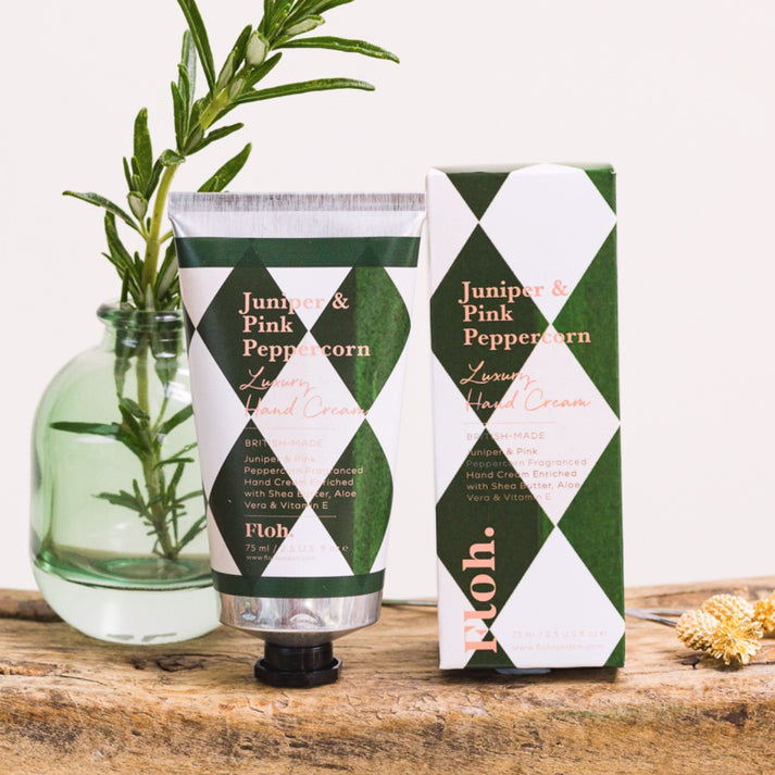 Toasted Crumpet Juniper & Pink Peppercorn Luxury Hand Cream - Floh. Juniper & Pink Peppercorn Lavender Hand Cream shown as a squeeze tube next to its retail box, styled on a rustic wooden surface with a green glass vase containing rosemary sprigs.