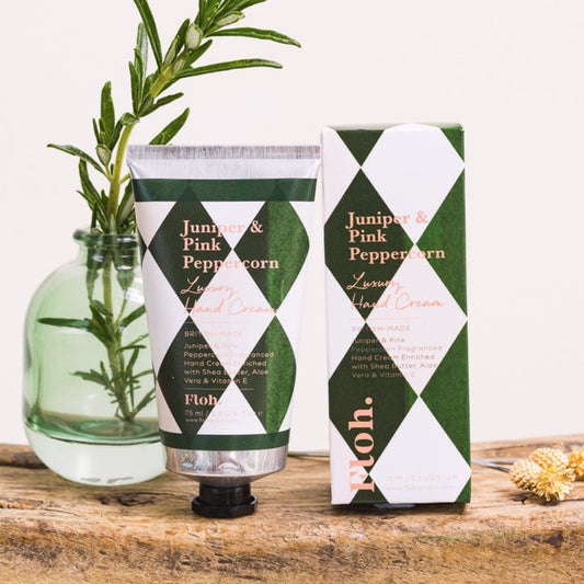 Toasted Crumpet Juniper & Pink Peppercorn Luxury Hand Cream - Floh. Juniper & Pink Peppercorn Lavender Hand Cream shown as a squeeze tube next to its retail box, styled on a rustic wooden surface with a green glass vase containing rosemary sprigs.