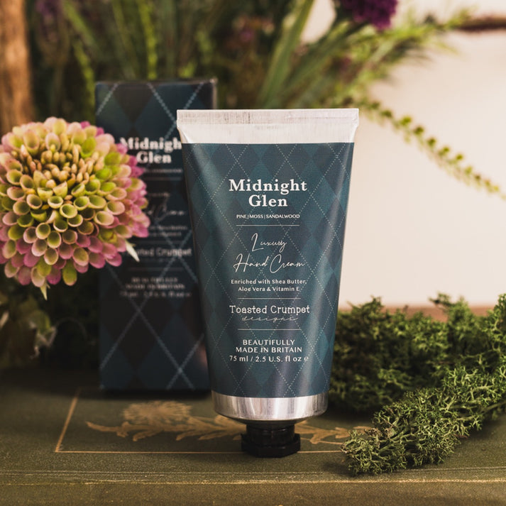 Toasted Crumpet Midnight Glen Luxury Hand Cream - A squeeze tube of Midnight Glen Luxury Hand Cream in Toasted Crumpet scent, standing upright on a rustic wooden surface with its box behind it and flowers and greenery around it.
