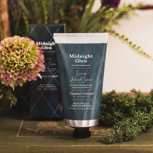 Toasted Crumpet Midnight Glen Luxury Hand Cream - A squeeze tube of Midnight Glen Luxury Hand Cream in Toasted Crumpet scent, standing upright on a rustic wooden surface with its box behind it and flowers and greenery around it.