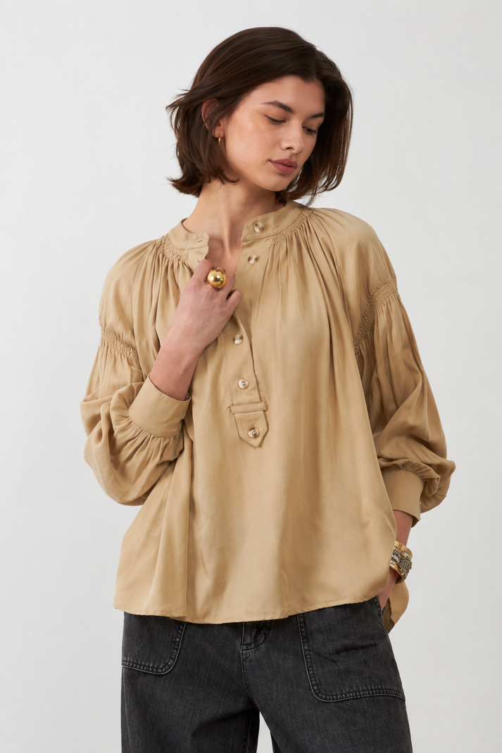 Orfeo Abby Shirt in Beige - a woman wearing a beige long-sleeve blouse with a gathered, smocked bodice and button placket, paired with dark grey jeans, photographed against a light grey studio background.