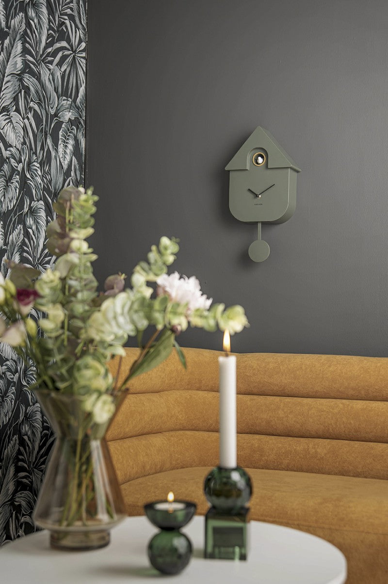 Green cuckoo clock on a gray wall with a vase of flowers, candle, and green vase on a table.