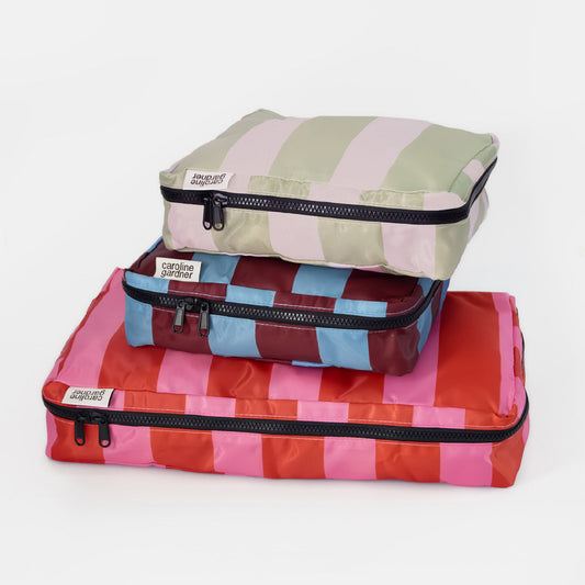 Three stacked zippered packing cubes from Caroline Jardin on a clean white background. The bottom cube is red and pink striped, the middle is blue and burgundy striped, and the top is sage green and pink striped.