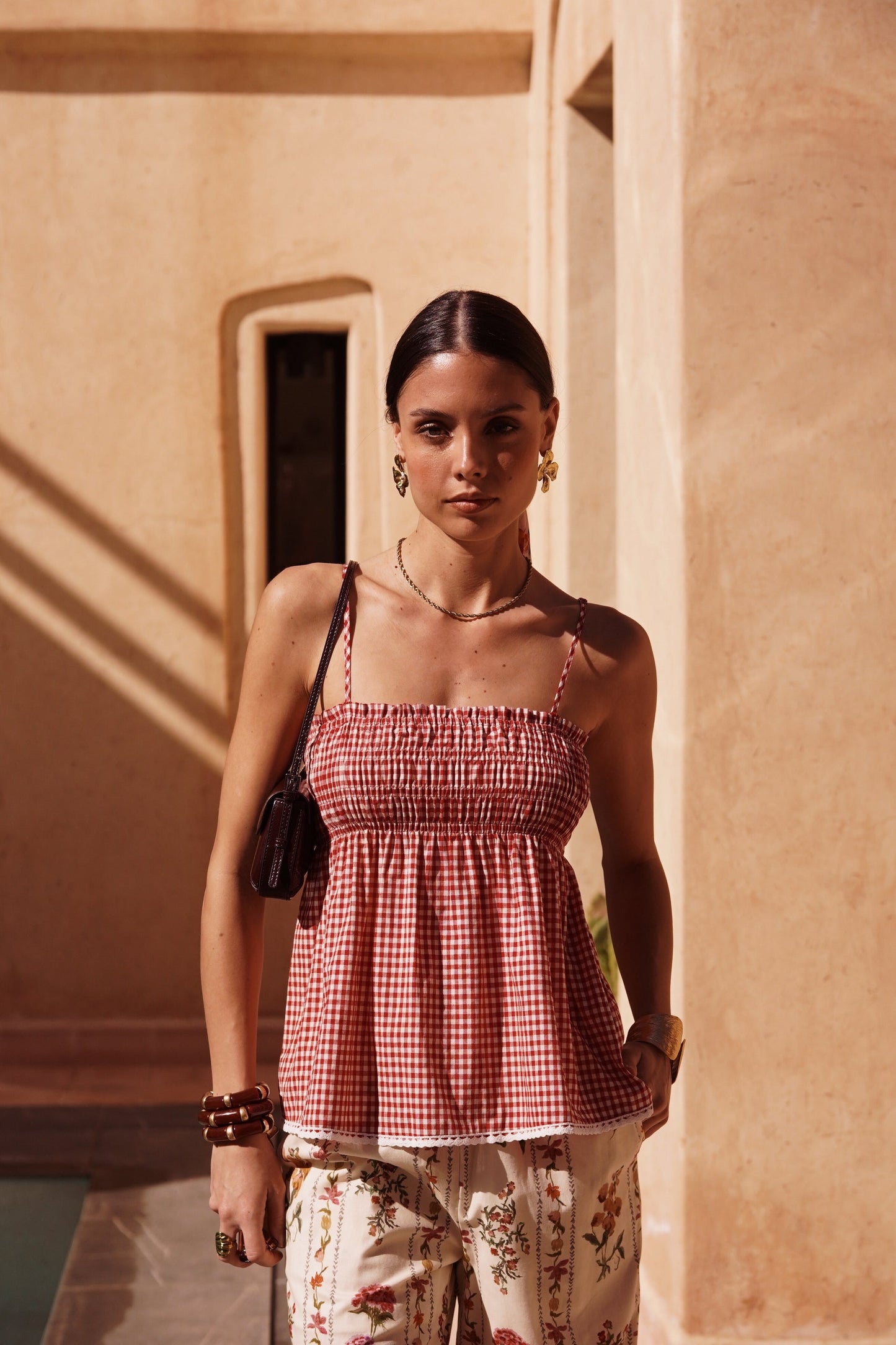 Woman in a red checkered top and floral pants standing in front of a beige wall.