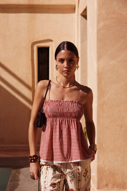 Woman in a red checkered top and floral pants standing in front of a beige wall.