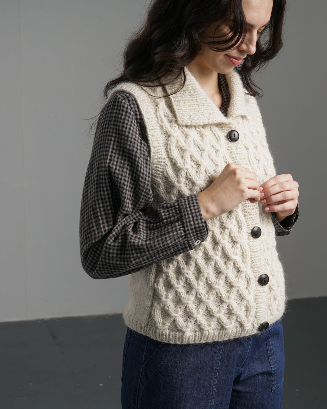 Woman wearing a cream knitted vest over a plaid shirt against a gray background