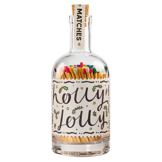 A clear glass bottle with a cork stopper, filled with colorful matches. The bottle has a white label with 'holly jolly' written in black script, decorated with small green holly leaves and gold stars.