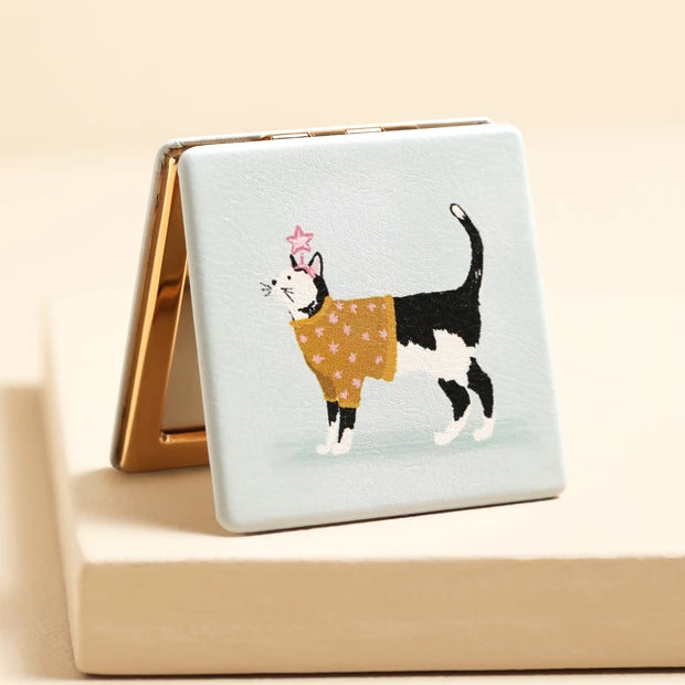 A square compact mirror with a light blue cover featuring an illustration of a black-and-white cat wearing a mustard yellow flower-patterned sweater, standing on a beige surface.