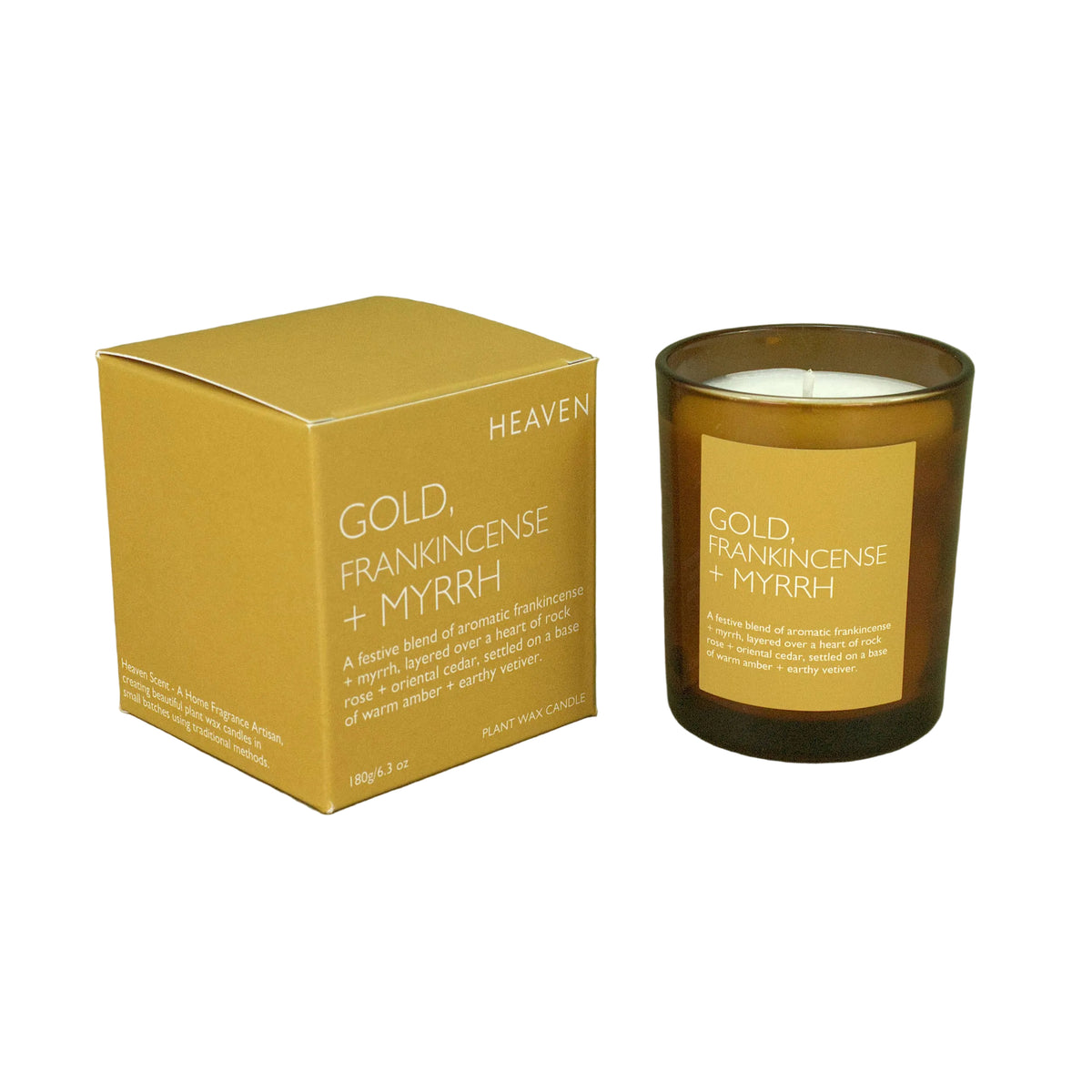 A candle in an amber glass jar next to its matching gold box, labeled 'HEAVEN GOLD, FRANKINCENSE + MYRRH', set against a solid olive green background.