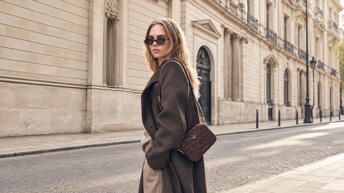 A woman wearing a dark brown trench coat and sunglasses stands on a city street next to a grand beige stone building, carrying a brown Leopard print shoulder bag by Katie Loxton