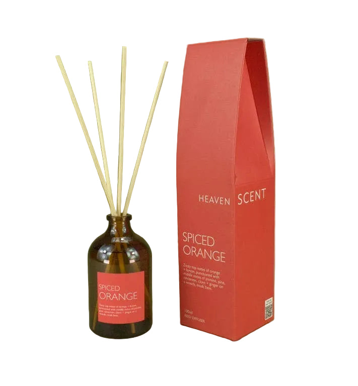 Amber glass reed diffuser bottle with four natural reeds and a coral-red label, standing next to its tall red packaging box on an olive green background. The box reads 'HEAVEN SCENT' and 'SPICED ORANGE'.