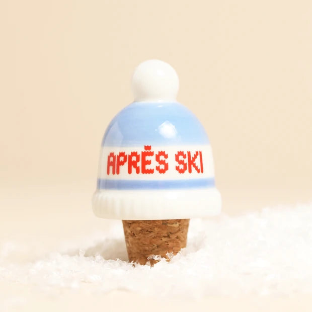A miniature cork bottle stopper shaped like a knit beanie hat, featuring a white pom-pom, light blue and white stripes, and the red text 'APRÈS SKI' on the front, set in white faux snow against a warm beige background.