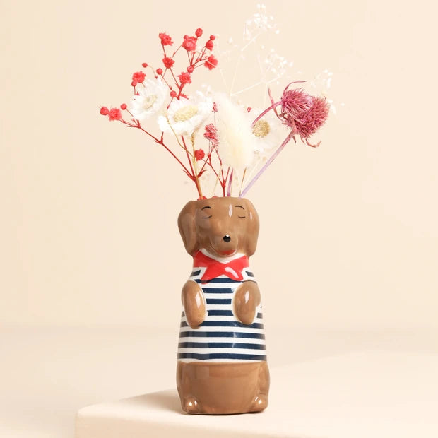 A ceramic dachshund-shaped vase with a navy and white striped body, a red bow tie, and closed-eyes detail, holding dried flowers against a warm beige background.