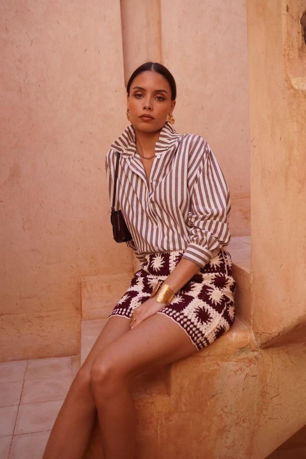 Woman sitting on a stone ledge wearing a striped shirt with frill neck and patterned skirt.