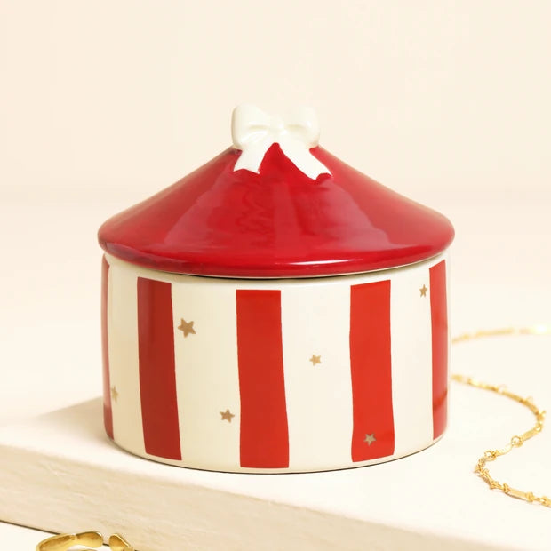 A round ceramic jewelry box with a red and white striped body, a glossy red lid, and a small white bow on top, styled on a light surface with a gold chain nearby.
