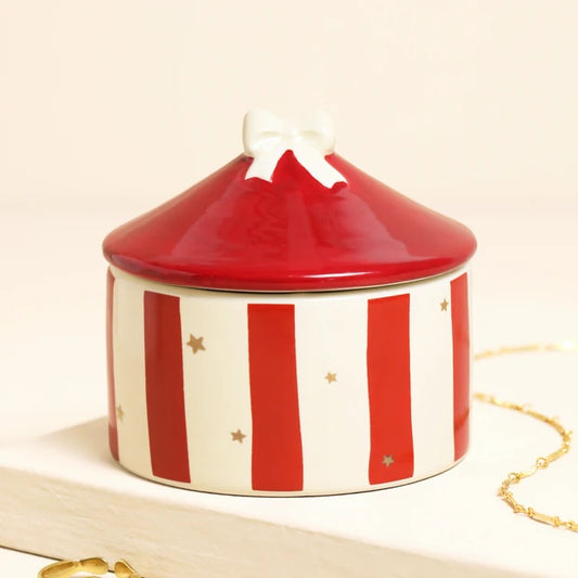 A round ceramic jewelry box with a red and white striped body, a glossy red lid, and a small white bow on top, styled on a light surface with a gold chain nearby.