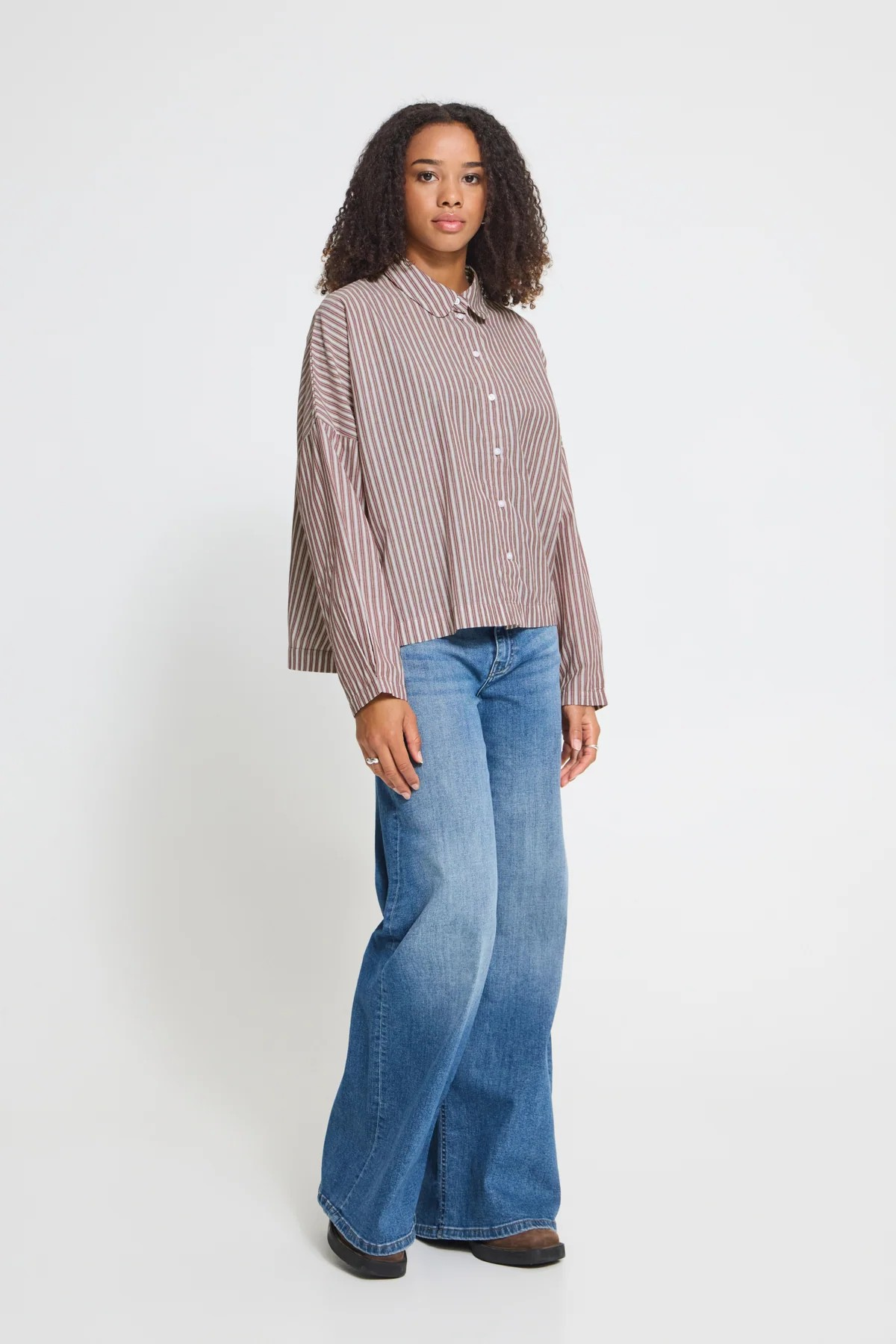 A woman modeling a long-sleeve button-up shirt with thin vertical stripes in brown and white, paired with wide-leg blue jeans and dark brown shoes, against a plain light gray studio background.