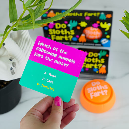 Colorful card with a trivia question about animals farting, held by a hand, with a book and small round object in the background.